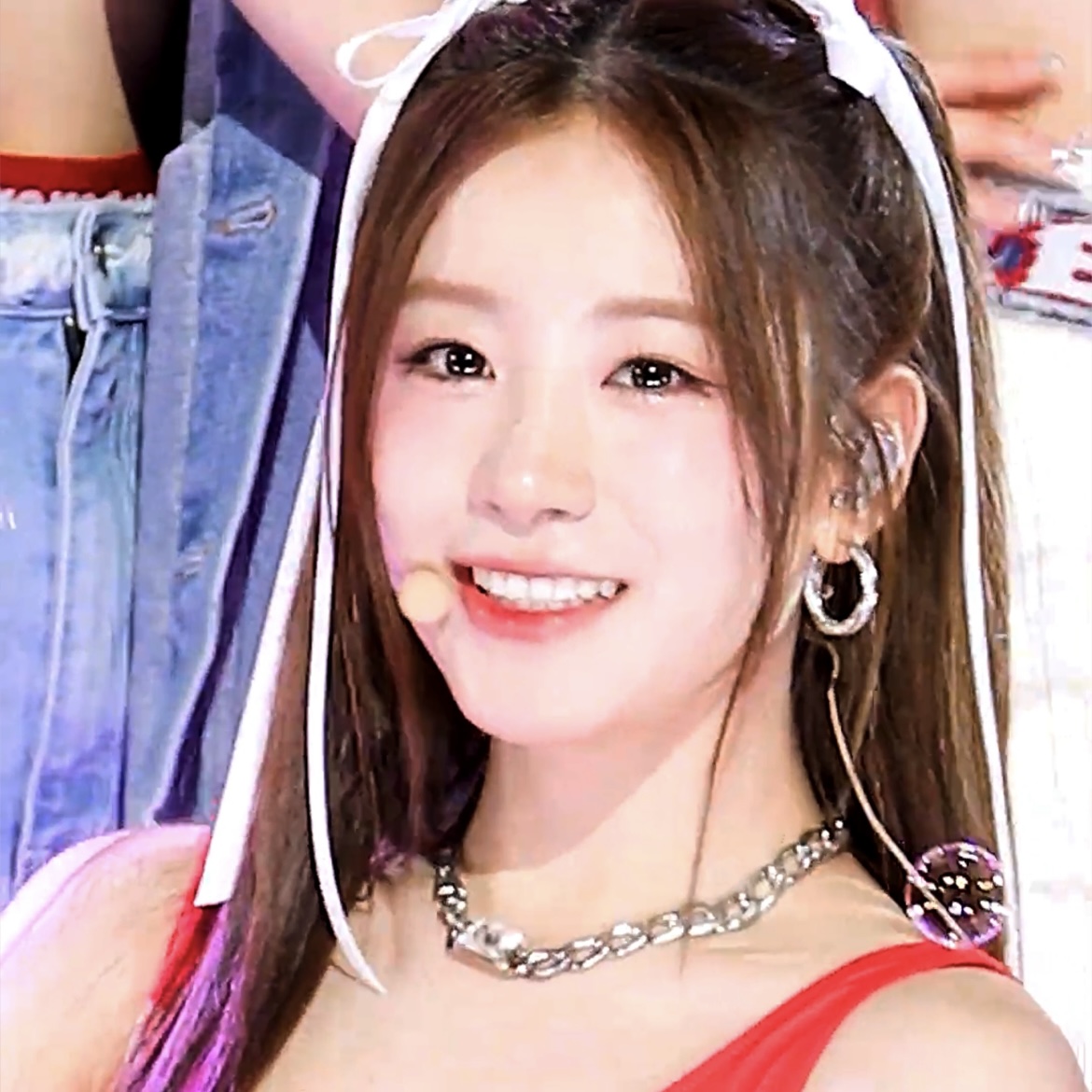 A close-up of a smiling young woman, likely a performer, wearing a headset microphone, a silver necklace, and a red top.