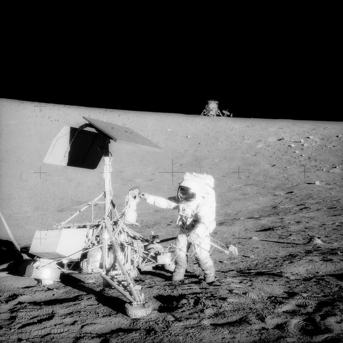 In this black and white photo taken on November 20, 1969, Apollo 12 Commander Pete Conrad stands with NASA's Surveyor 3 lander on the dusty surface of the Moon. The Apollo 12 Lunar Module Intrepid is visible on the horizon in the background.