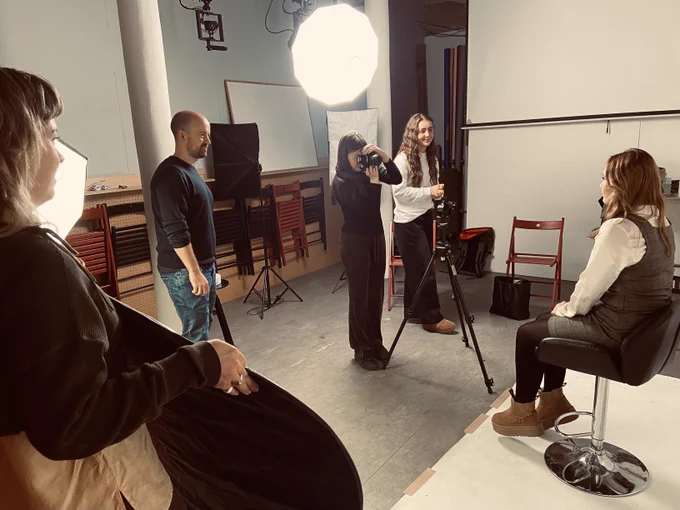 A young person is sitting on a chair in a studio, being photographed by a student with a camera and three teammates.