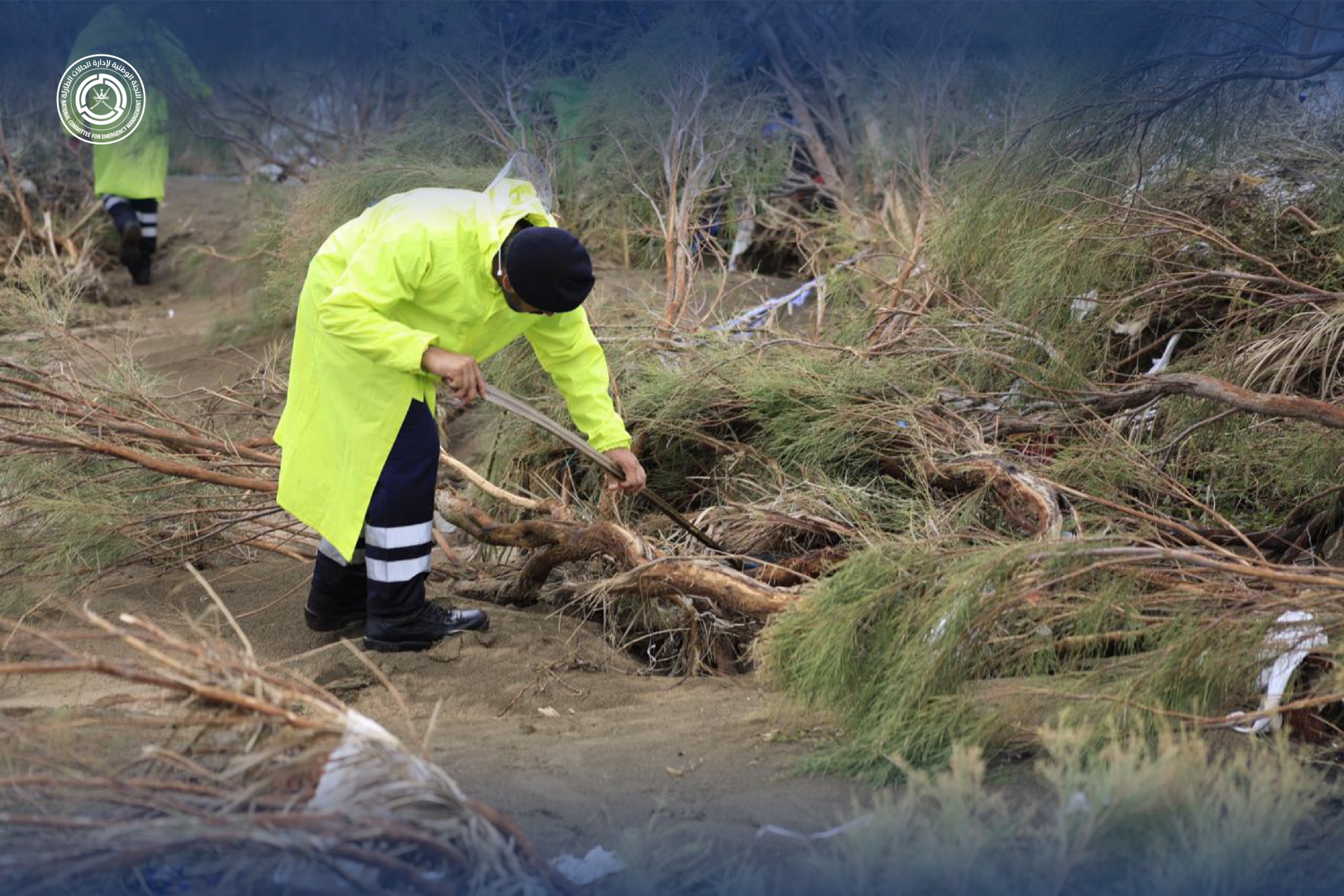 فرد من أفراد شرطة عمان السلطانية يرتدي سترة ضد المطر ممسكا بعصا طويلة ومنحنيا إلى الأسفل حيث الأشجار الصغيرة الموجودة على جانب الوادي بحثا عن المفقودين 