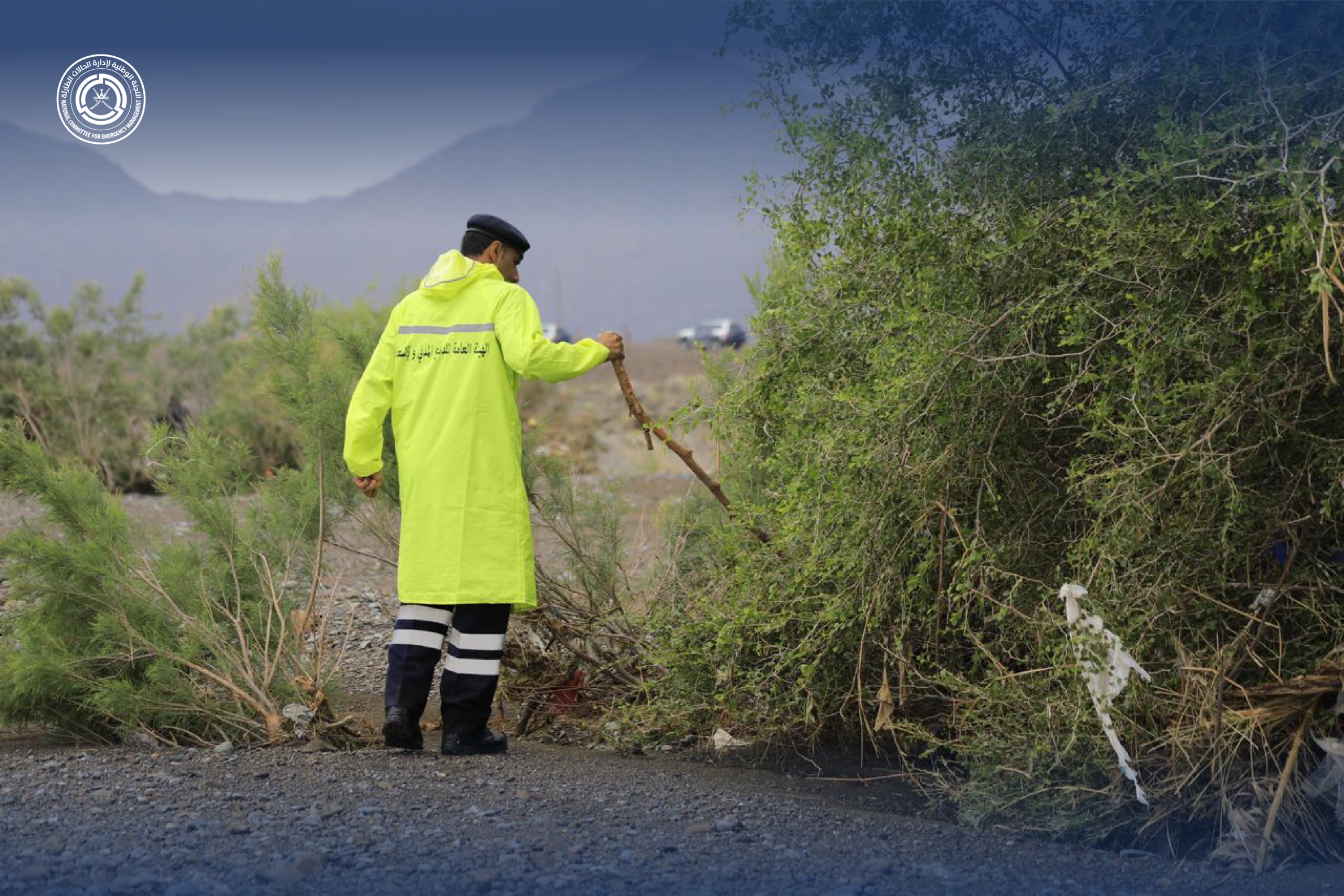 فرد من أفراد شرطة عمان السلطانية يرتدي سترة ضد المطر ويحمل في يده اليمين عصا كبيرة يفتش بها ما تحت الأشجار الظاهرة بالوادي بحثا عن المفقودين