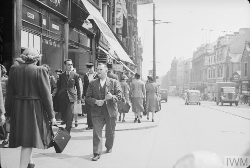 Men and women go about their daily business on this busy street in Newcastle-upon-Tyne. Several vehicles can also be seen. You can see more photos of life on the home front on our website: bit.ly/3TF90gd © IWM (D 15659)