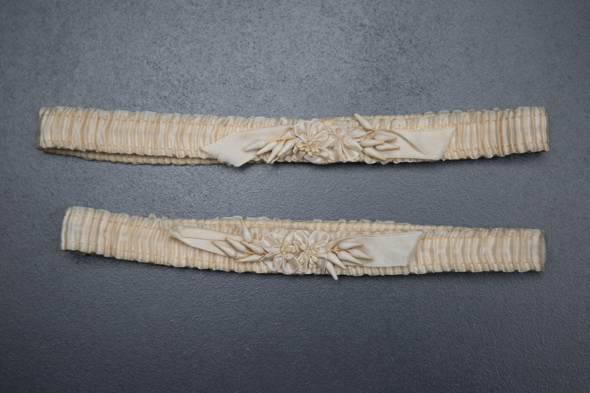 This pair of garters has an elastic base, topped with an ivory silk ribbon, and is embellished with silk ribbonwork flowers and wax floral stamens. They were likely intended as bridalwear for single use, as the wax would not have held up to long-term wear or washing.