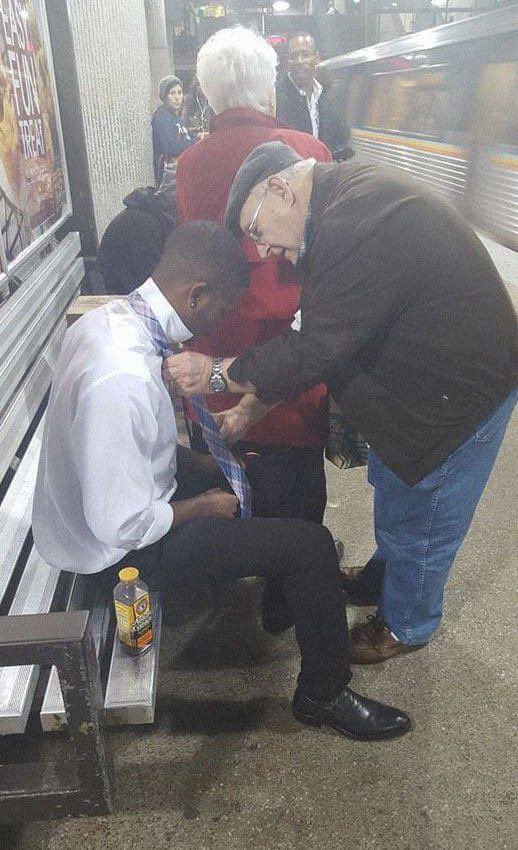 The young guy sitting down was struggling with his tie. The woman in the red coat noticed, and asked ‘Do you know how to tie it properly?’ The young guy said ‘No ma’am.’ She taps her husband and says ‘Come to this side and teach this young man how to tie his tie.”