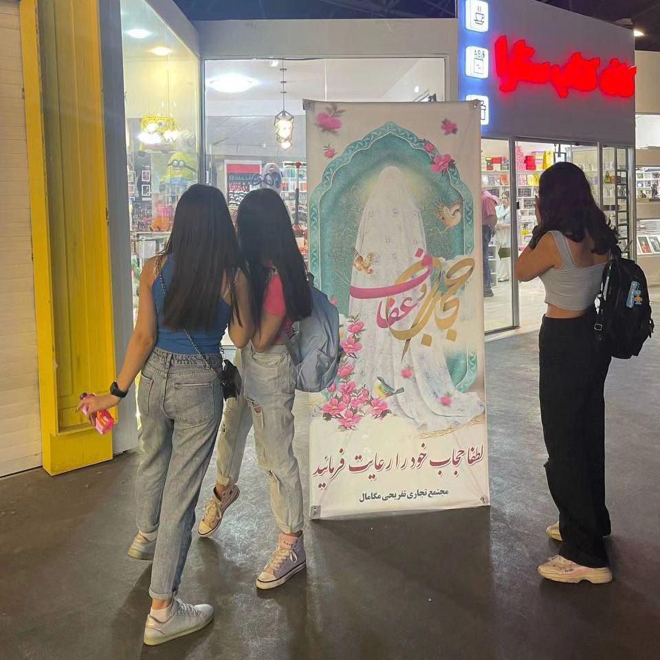 Iranian girls in Iran pose without hijabs beside a state hijab advertising banner. One way or another, they will win this fight.