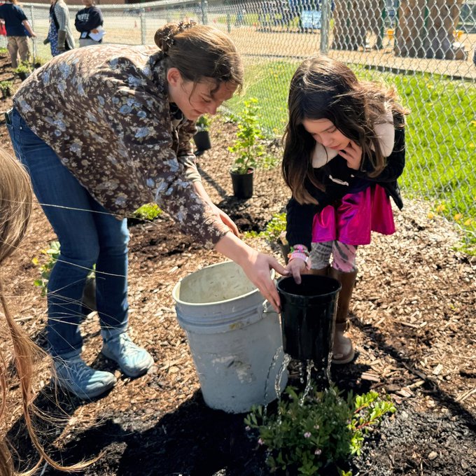 A high school student helps an elementary school student with the planting process, working with a bucket and planter bucket