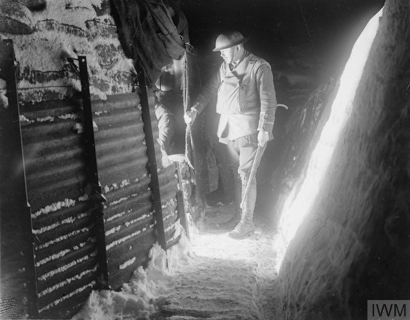 An officer of the 12th Battalion, East Yorkshire Regiment, draws the gas curtain of a dug-out - in this atmospheric image by 2nd Lieutenant Thomas Keith Aitken - while on his rounds in a snow-covered support trench near Roclincourt, 9 January 1918. IWM (Q 10621)