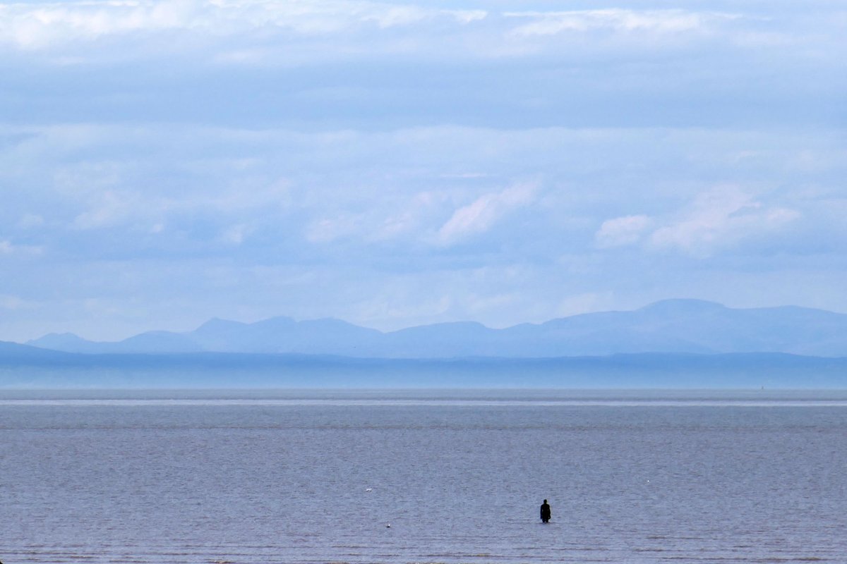 A fine start to the Sunday of the Bank Holiday weekend, so many shades of blue down on Crosby beach today. The sea calm and a light sea mist rising along the distant Welsh coast, the mountains of Eryri, Snowdonia, clear of any cloud this morning. @IronMenCrosby