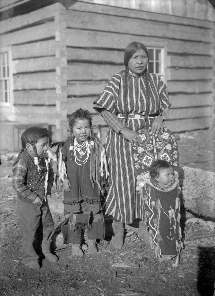 Lucy, a Native American woman on the Flathead Indian Reservation in western Montana, stands with three children in front of a wood building. Her hair is in long braids; she wears a striped dress with a beaded belt. She holds a cradleboard with a baby; the boys have spiked hair.