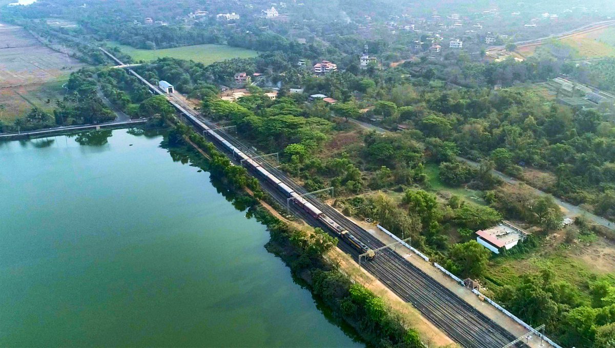 A freight train passes by the expansive stillness of a lake near Karmali Station, Goa fringed with green wilderness.