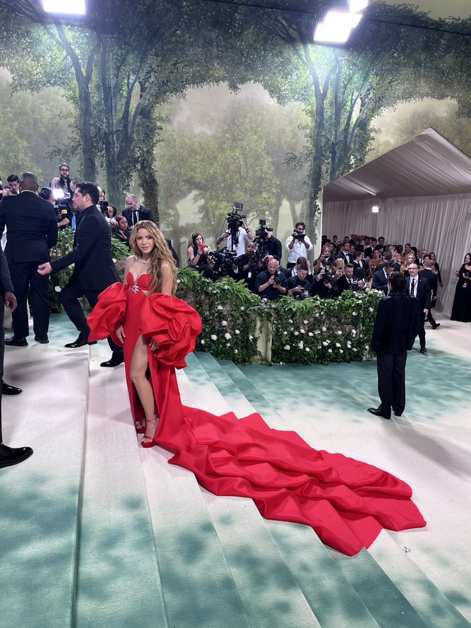 Shakira poses on the stairs of The Met Gala red carpet in a red gown. The large train of her dress drapes on the stairs below her. 