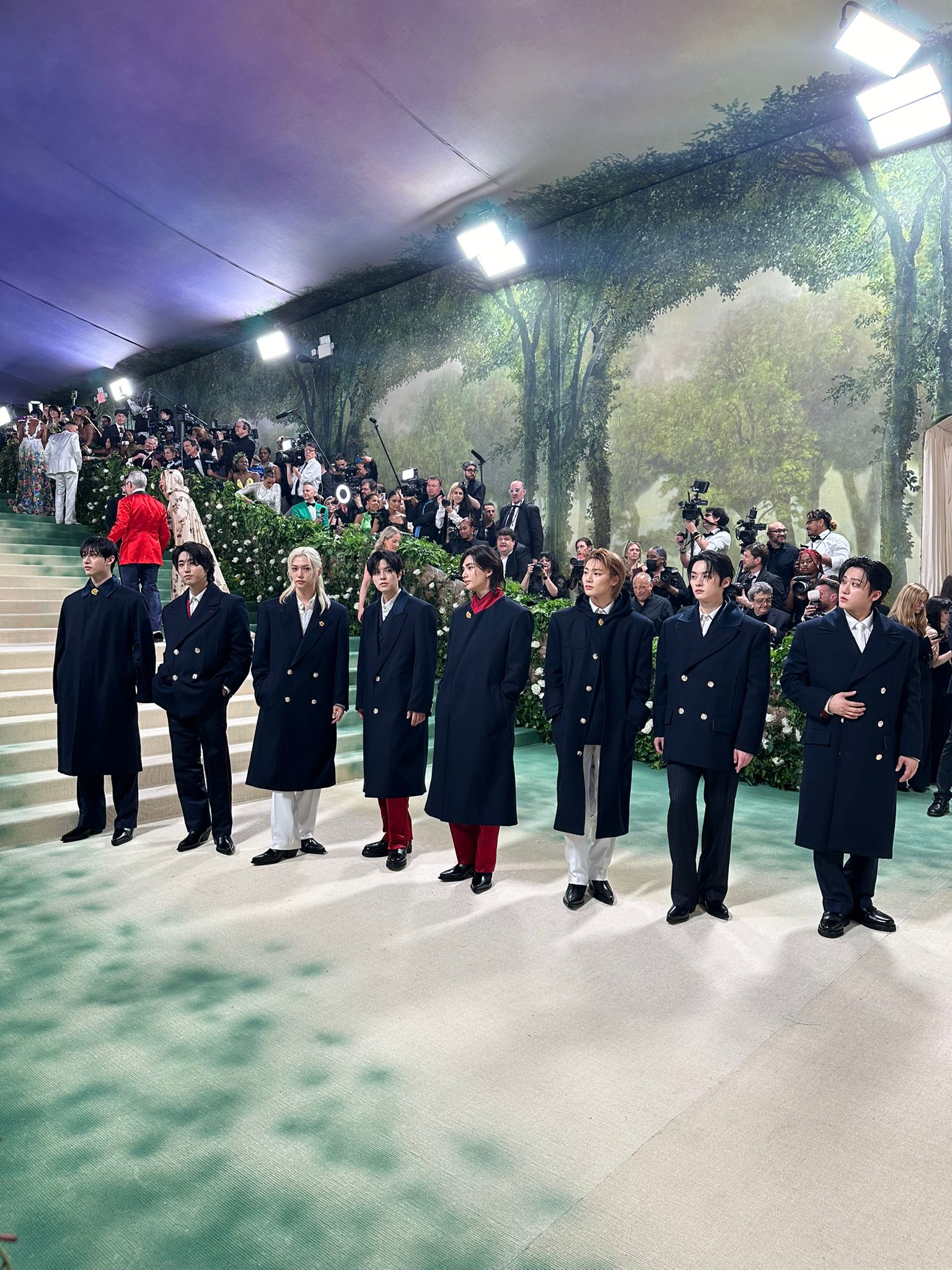 The eight members of the boy band Stray Kids pose together in a line on The Met Gala red carpet. They're wearing coordinating navy blue jackets. 
