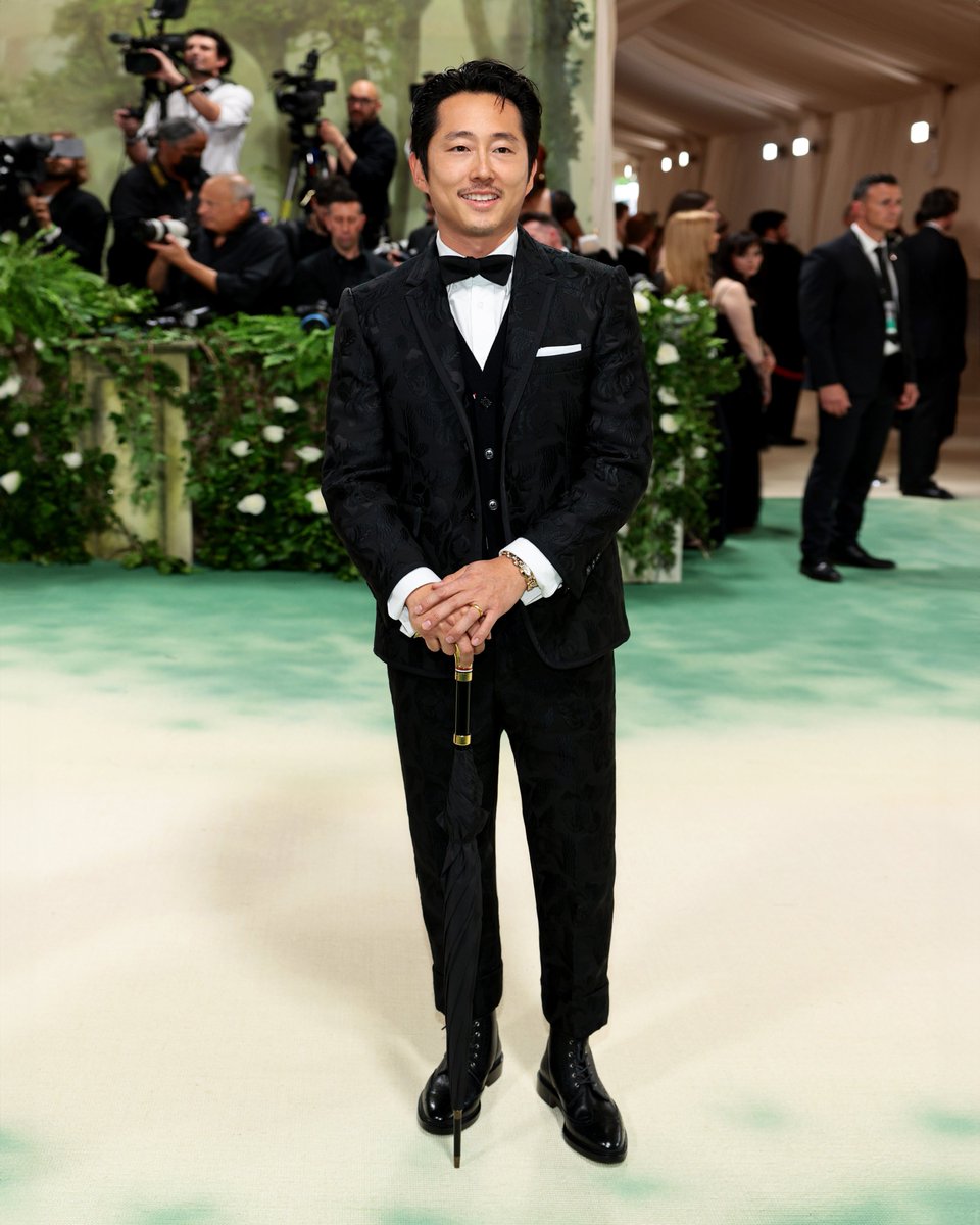 … steven …
steven yeun wears a made-to-measure thom browne tuxedo in black roses and ravens tonal jacquard with black grosgrain tipping over a cable knit vest in black cashmere, blucher cut tuxedo shoes in black patent leather and umbrella with gold raven handle for the 2024…