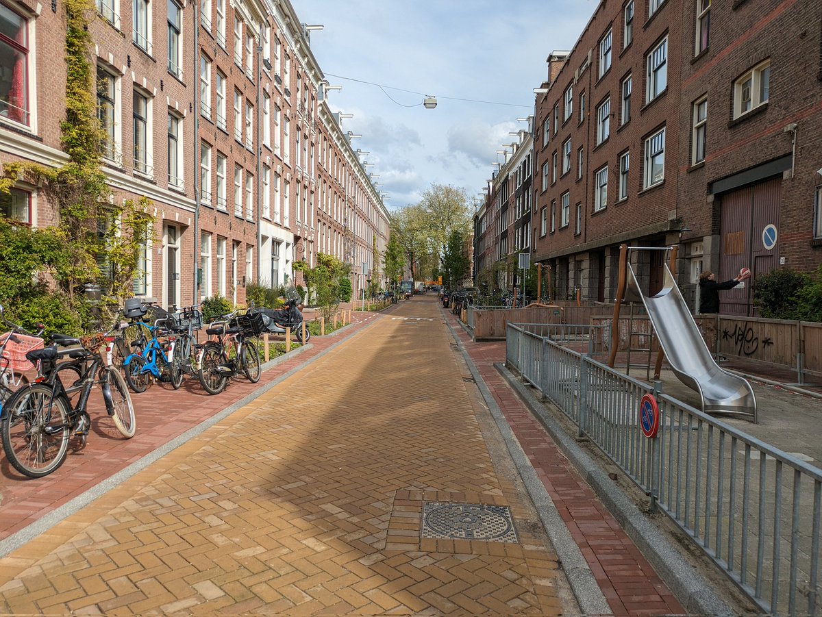 For a few decades these residential streets were busy rat runs and most space was crammed with parked cars. Now all through motor traffic is diverted elsewhere and most of the space is now used for greenery, cycle parking and a playground