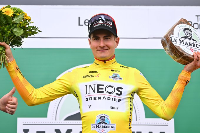 Carlos Rodriguez stands atop the podium in the yellow leader's jersey at Tour de Romandie with a bouquet of flowers in one hand and a large  slab of Swiss 'Le Marechal' cheese in the other hand. 