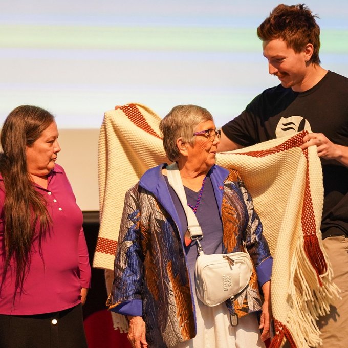 Wife of late former state senator John McCoy receives a gift from event hosts as a blanket is wrapped around her on stage