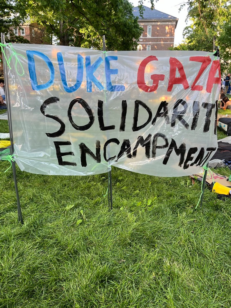 Photo of a banner: "Duke Gaza Solidarity Encampment" staked into grass, with people behind in the encampment at UNC-Chapel-Hill