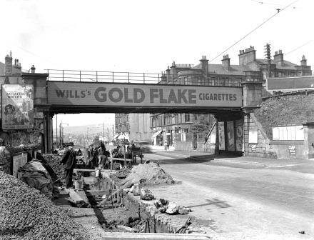 Hoarding on the old railway bridge at 1551 Great Western Road, Anniesland in August 1936. Workmen are working in an open trench parallel to the main road. Archive ref: D-CA8/1410