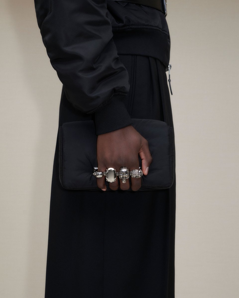 A flight jacket and cigarette trousers in black, worn with a racer-back harness and a nylon puffy #McQueenKnuckle clutch. Discover the #McQueenMenPreAW24 collection: on.alexandermcqueen.com/PreAW24Mens_TW