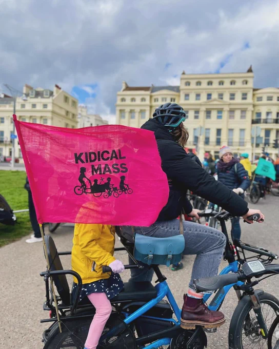 Lady on e-cargo bike with a girl on the back and a large Kidical Mass flag. Kidical Mass ride April 2024 - Hove and Brighton. Photo by Aaron Fegan