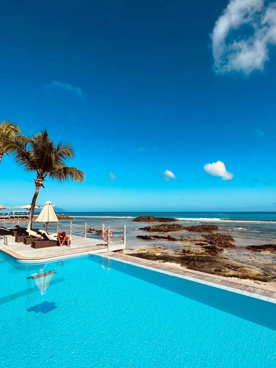 An idyllic escape where azure skies kiss tranquil sea blues, a solitary palm sways, and the sun basks in serene warmth. The pool, a mirror to the heavens, cradles a couple's secluded paradise by the whispering waves.