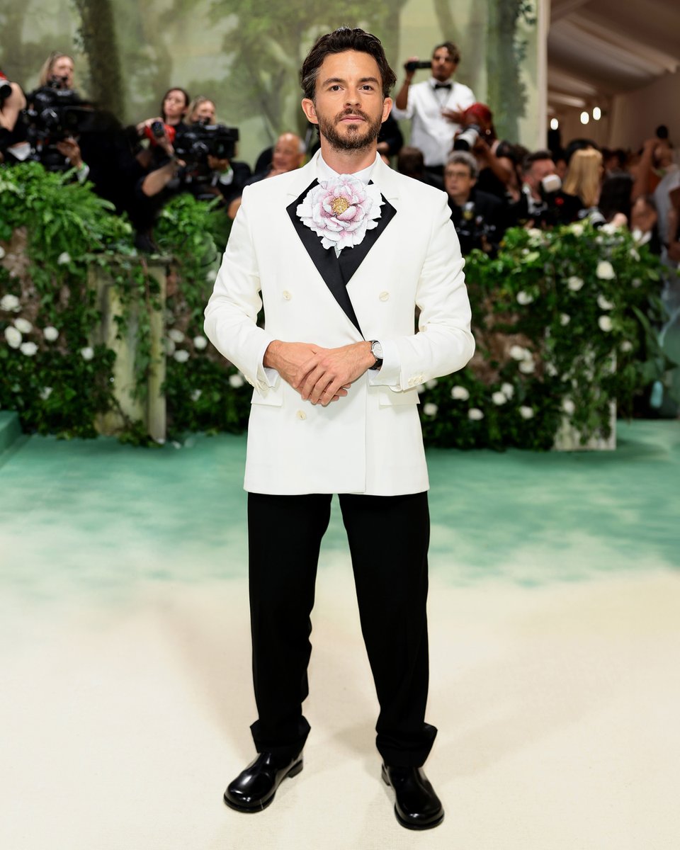 Jonathan Bailey in bespoke LOEWE at the Met Gala 2024. A double-breasted white jacket with a black satin lapel is paired with classic black tailored trousers. The shirt features a handcrafted peony flower; each metal leaf is carefully shaped and hand painted and embellished with…