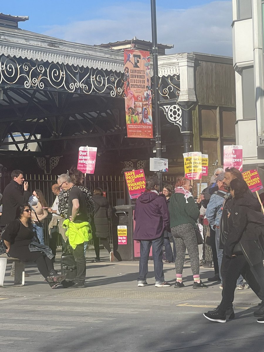 They shout “Say it loud, say it clear, refugees are welcome here.” He’s yelling out that migrants are coming from the poorest places in the world- “desperate people fleeing torture and war….” Most of the group have white hair or bald heads. They lived most of their lives in a