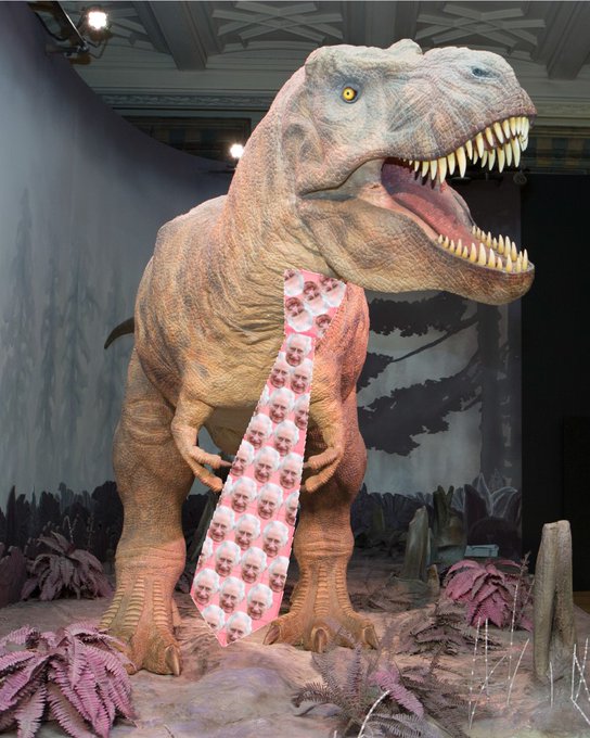 A photo of the animatronic T. rex at the Natural History Museum wearing a photoshopped pink tie, patterned all over with King Charles' face.