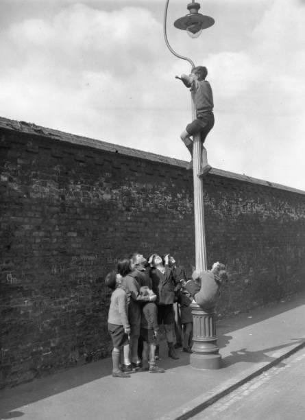 A photograph of a boy watching a cricket match at the Oval, London, from the top of a lamppost, while his friends wait below for a commentary. Taken in 1938.