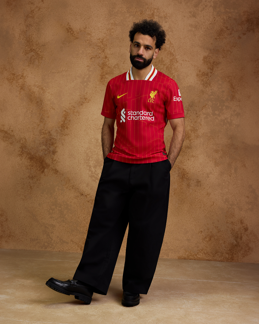 Mo Salah poses in our new 2024/25 home kit. The red jersey features a chrome yellow pattern design which sees a modernised take on the legendary '84 home shirt, a season which saw the Reds become European Champions for the 4th time in Rome and the first English Club to win a Treble of major honours.