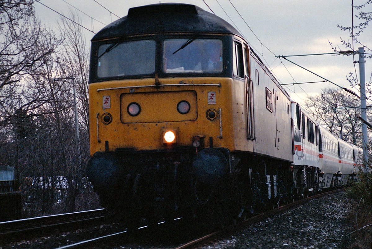 POYNTON: 47537
A cold winter Sunday sees 'Sir Gwynedd - County of Gwynedd' dragging a class 90 north to Manchester on a down Euston service. Quite a dramatic angle through a fence, but straight into the sun. Winter 1990.