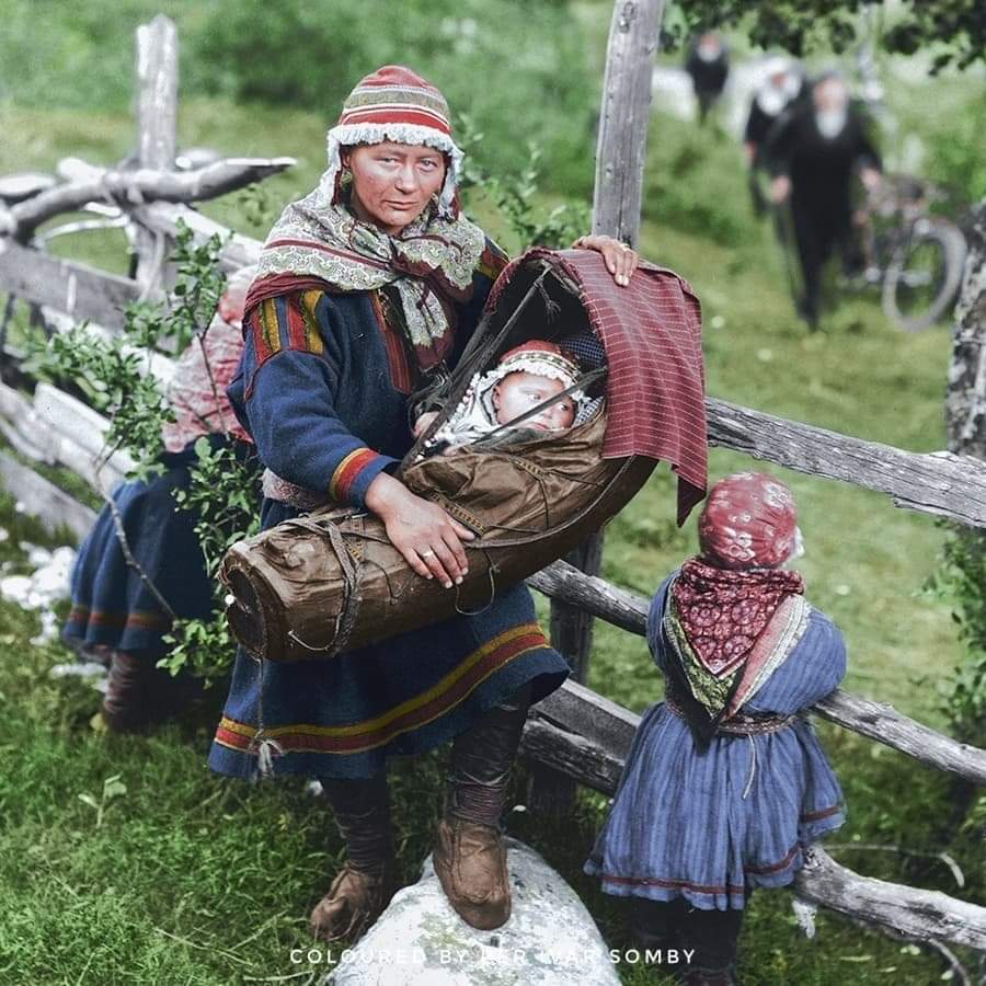 A Sami woman photographed standing by a wooden fence with her children in Lyngen, Troms, in c. 1925. Photograph taken by Anders Beer Wilse.