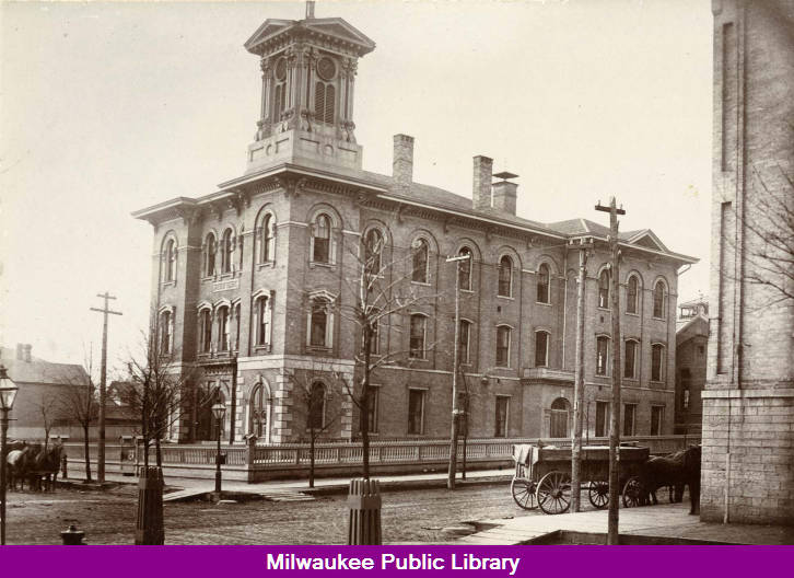 Humm! This photo shows an unidentified brick schoolhouse in Milwaukee. It is a multistory building with a clock tower. Horses and wagons can be seen beside the building.