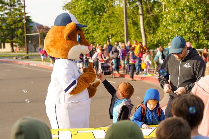 A student high-fives Dinger, the Bellingham Bells mascot, while a group of parents and students walk on sidewalk in background