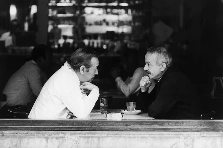 Roberto Goyeneche y Astor Piazzolla en un bar porteño en los años 80, charlando sentados a una mesa junto al ventanal.
