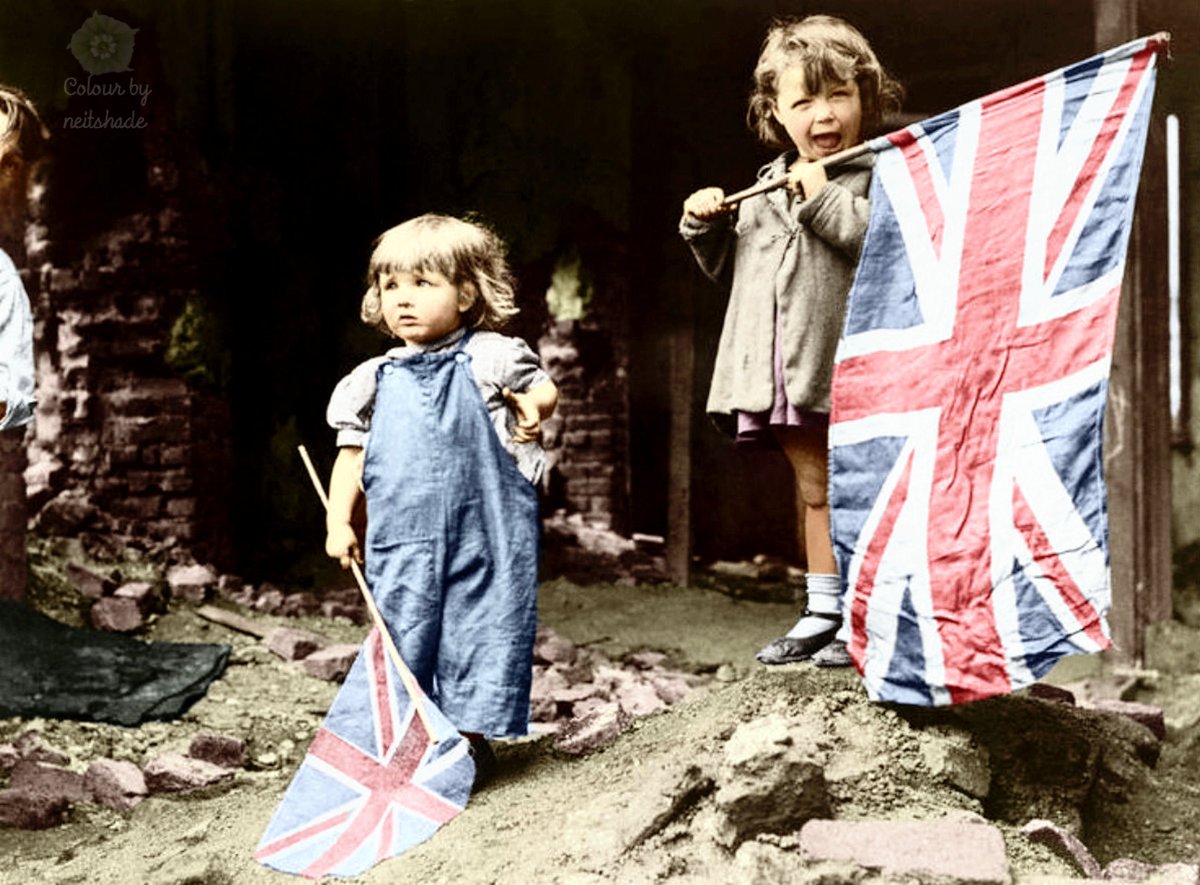 VE Day in London, 8th May 1945. Two small girls waving their flags in the rubble of Battersea, snapped by an anonymous American photographer. #VEDay