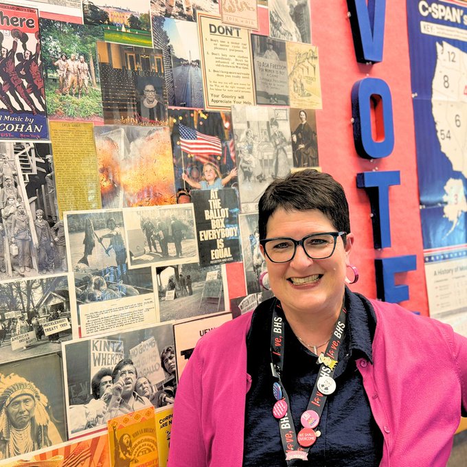 Jen Reidel poses for photo with wall sharing news clippings and images related to social studies and the the word VOTE in big letters on wall