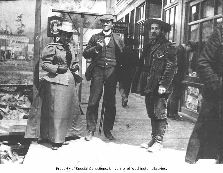 Circa 1900, a woman and two men standing on a wooden sidewalk in Skagway. This photo could launch an entire essay on fashion in gold rush era Alaska. Note the heavy skirt that could survive the mud and snow. #alaskahistory #skagway #alaska