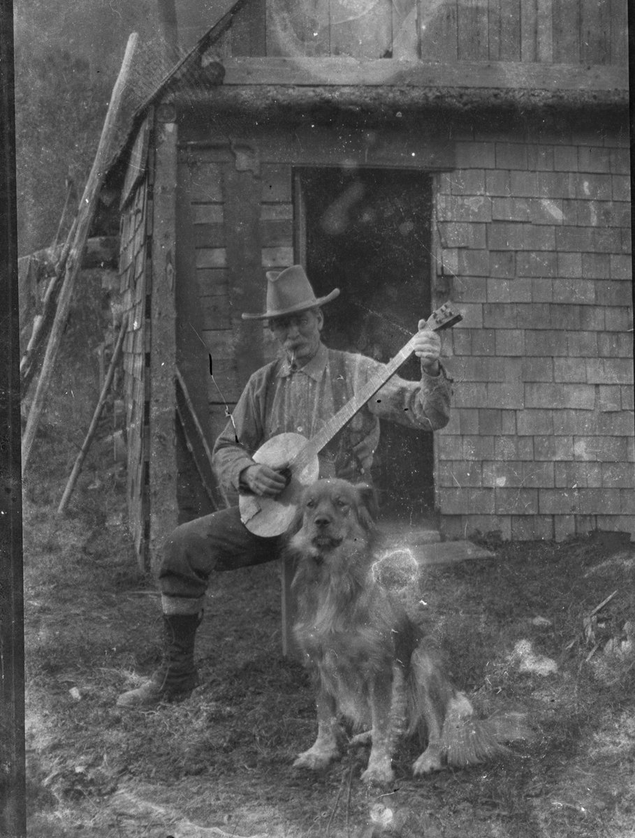 Circa 1910-1940 Alaskan man smoking a pipe and playing a banjo for his dog, likely around Alexander Creek, near the Susitna River mouth, northwest of Anchorage. Via Anchorage Museum. #alaskahistory #alaska