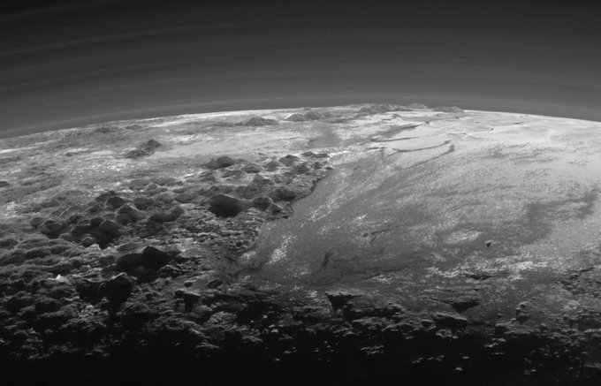 A grayscale image of Pluto’s landscape near sunset. A multitude of short, rugged mountains texture the surface. The icy plains appear as a spray of dark and light across the flat part of Pluto’s surface. Credit: NASA/JHUAPL/SwRI