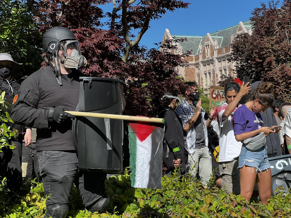 One of the encampment occupiers has an improvised shield made from a plastic garbage can. Also wearing a hammer and sickle patch.