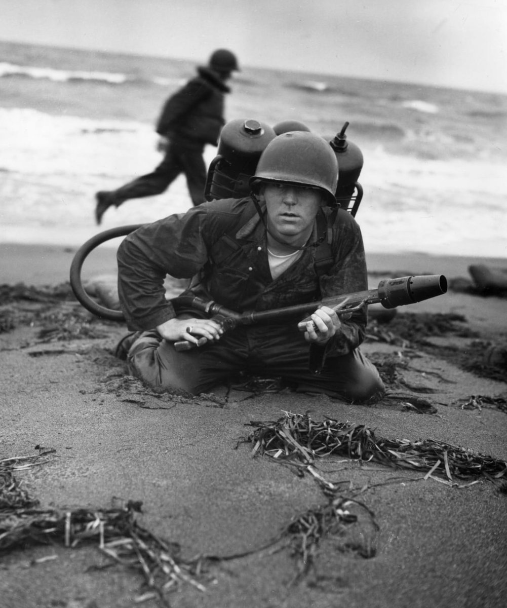 In 1945, a Marine crouches low to the beach, holding his flame-thrower during amphibious training at Chigasaki. 🪖