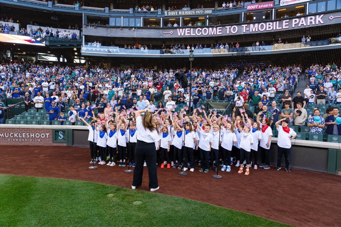 Center right view as students hold up flags on infield warning track while teacher conducts and fans look on. Photo courtesy of Ben VanHouten/Seattle Mariners
