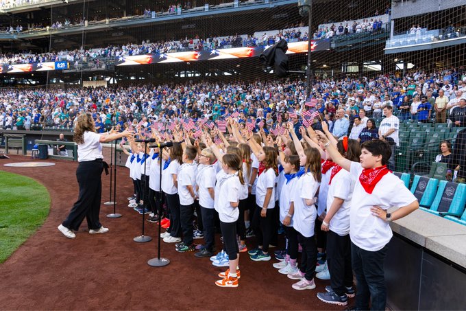 Right side view as students hold up flags on infield warning track while teacher conducts and fans look on. Photo courtesy of Ben VanHouten/Seattle Mariners