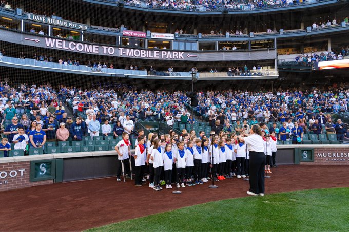 Center left view as students hold up flags on infield warning track while teacher conducts and fans look on. Photo courtesy of Ben VanHouten/Seattle Mariners