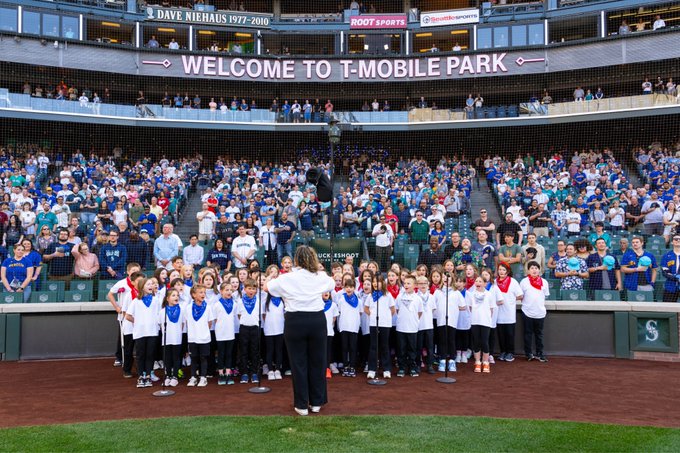 Straight on view as students hold up flags on infield warning track while teacher conducts and fans look on. Photo courtesy of Ben VanHouten/Seattle Mariners