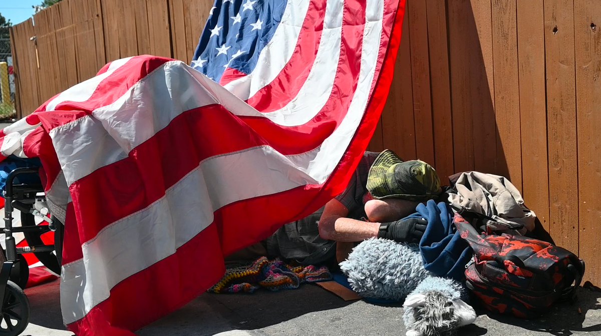 In photo, on the streets of Reno lying outside under the sun under a giant American flag, 65-year-old Don Newman, who says he's an Air Force veteran, says he has been unhoused for about a year now, after not having enough money to support himself and his wife.