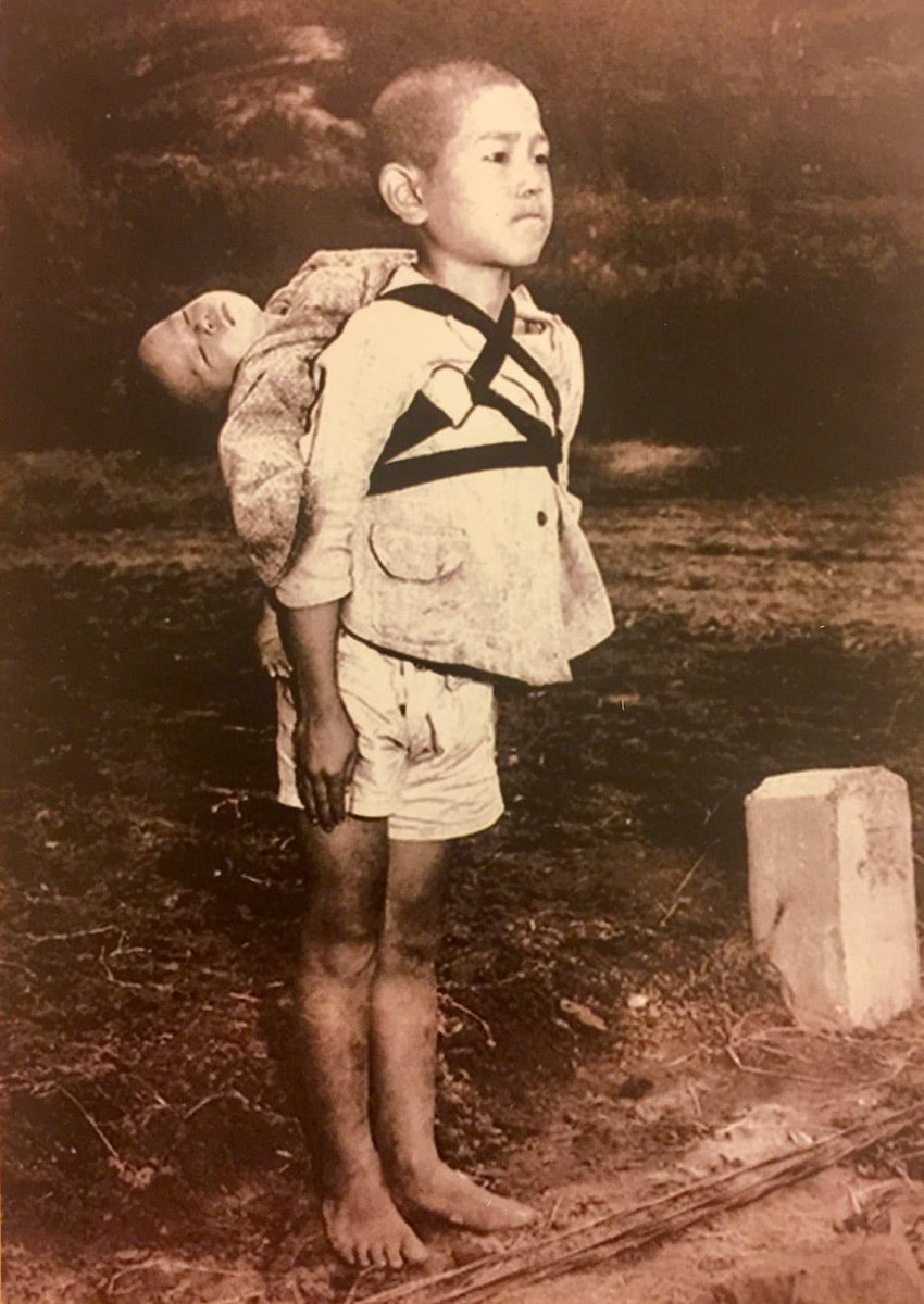 A Japanese boy waiting in line at the crematorium with his deceased baby brother on his back, Nagasaki, 1945.