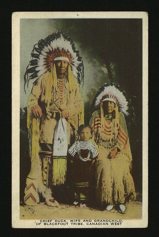 Hand-tinted photograph of Chief Duck, his wife and grandchild. All are dressed in ceremonial costume. Credit - Alberta News Ltd.: Publisher, Public domain, via Wikimedia Commons