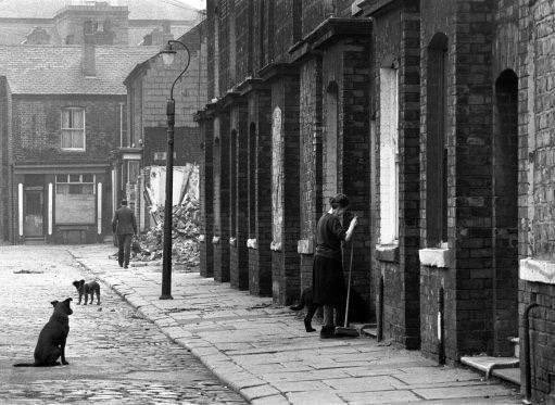 A photograph of a woman on a street Half demolished and the Neighbours all gone.   
In Lower Broughton still she kept the pavement clean in front of her house.